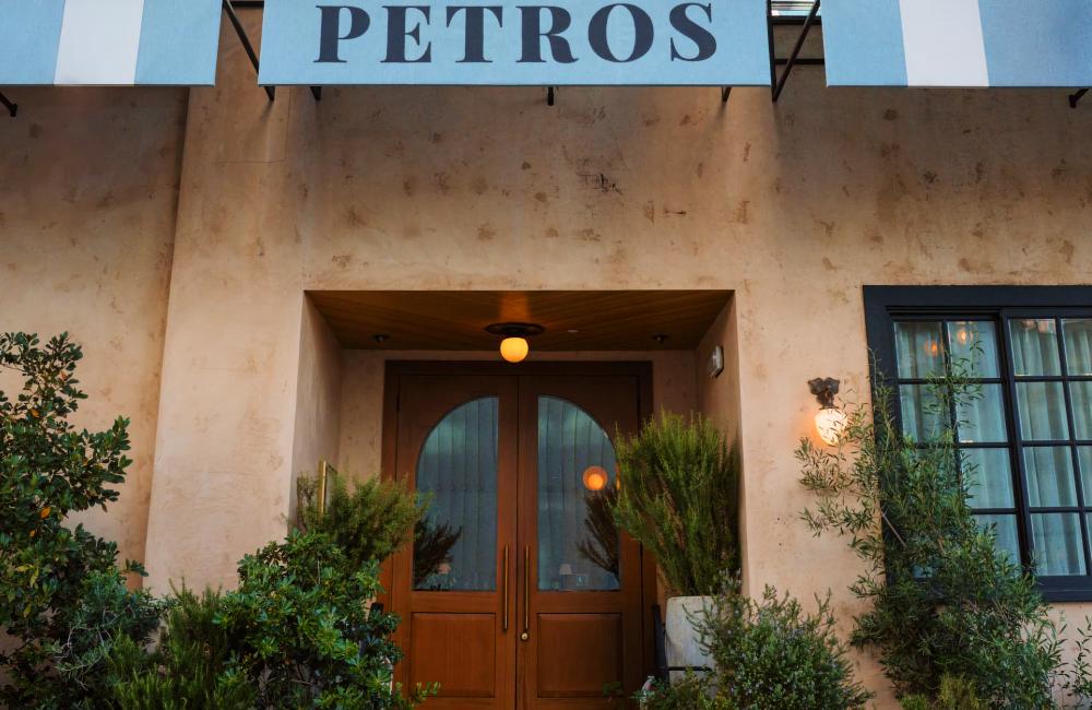 The entrance of Petros restaurant features wooden double doors, greenery in pots, and a blue-and-white striped awning.
