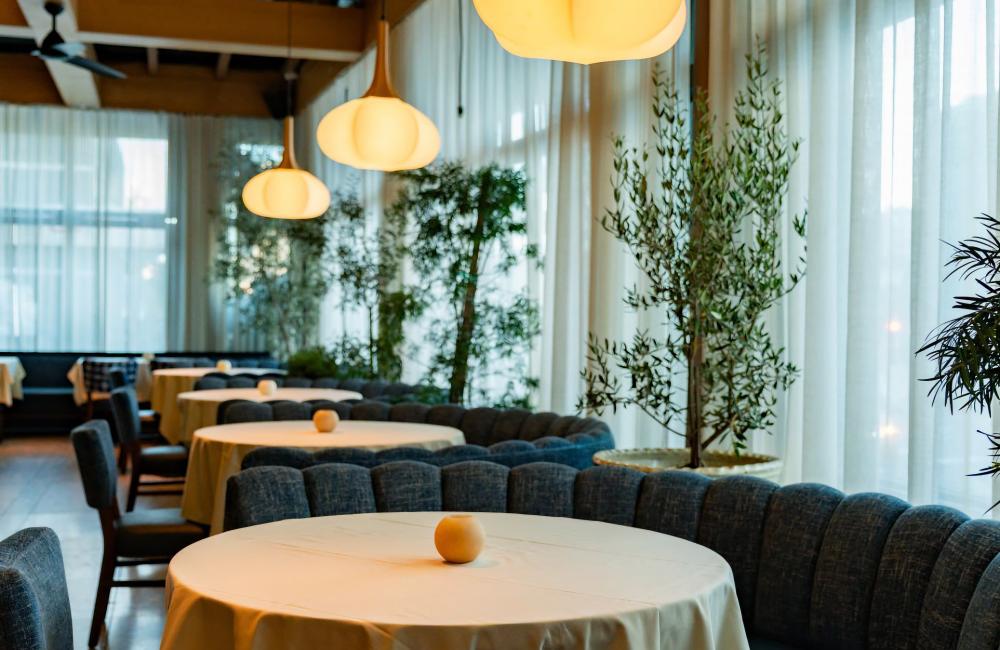 Round tables with white tablecloths and gray upholstered chairs line a softly lit restaurant interior with large windows and hanging pendant lights.