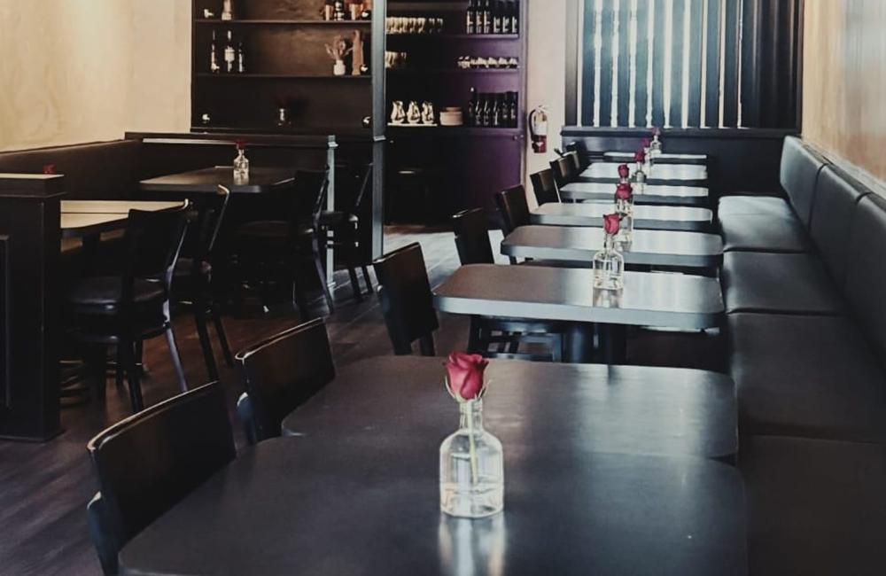 A restaurant interior with empty dark tables, each decorated with a single red rose in a glass vase; shelves with bottles and décor in the background.
