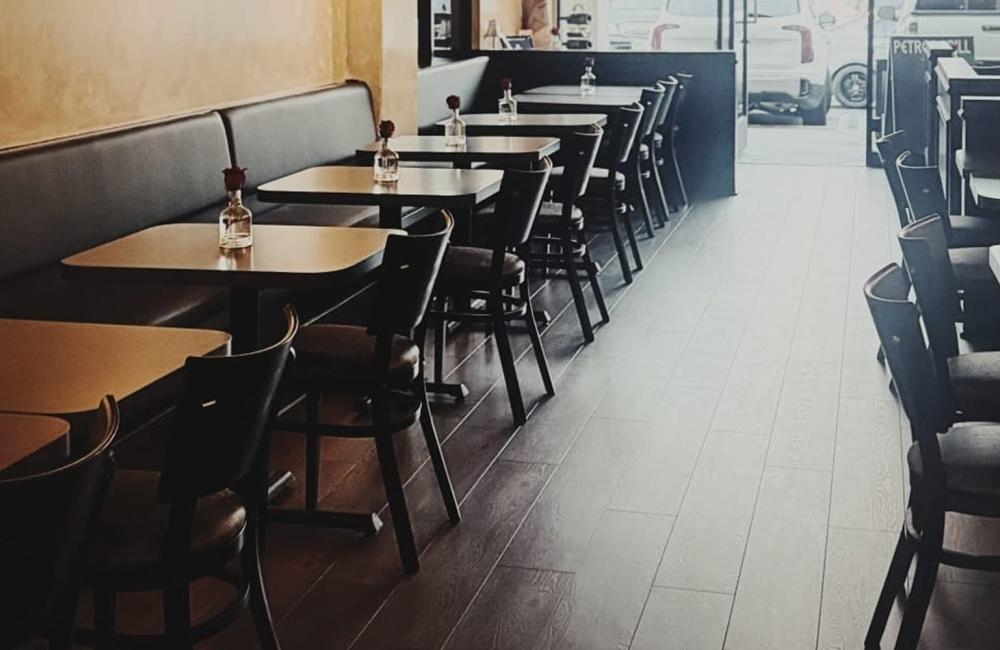 Empty restaurant with dark wooden tables and chairs, single flower vases on each table, and natural light coming through the front windows.