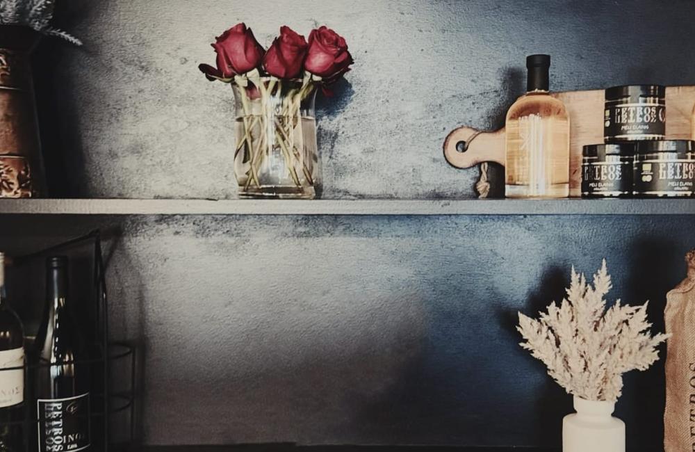 Two shelves display bottles of wine, a vase with dried flowers, skincare products, a cutting board, a small bouquet of red roses, and various decorative items against a dark background.