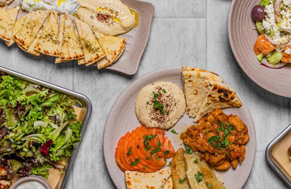 Several plates of Mediterranean food, including grilled skewers, pita bread, hummus, rice, salad, and vegetables, arranged on a light-colored tabletop.