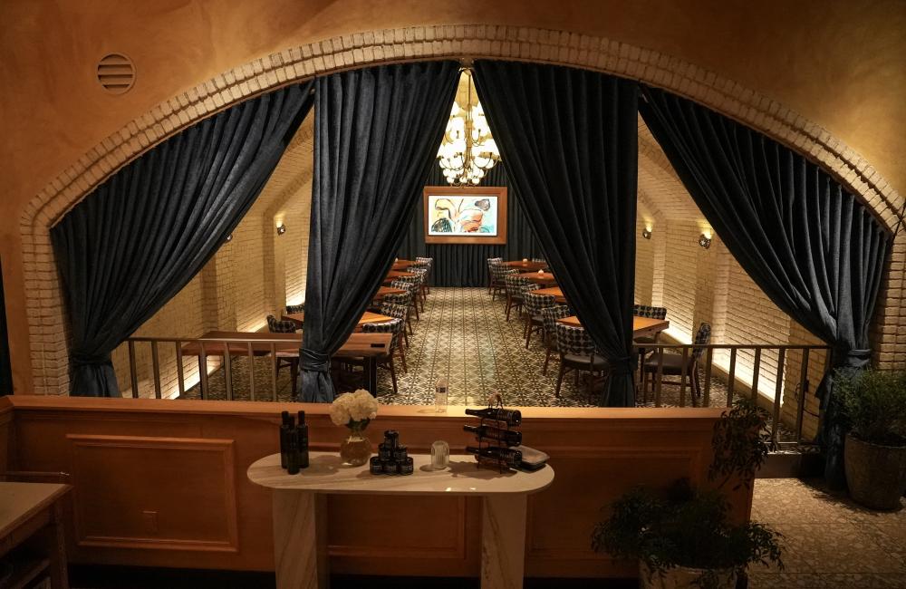 A dimly lit dining area with patterned floor tiles, tables, and sculpted blue curtains under an arched brick ceiling.