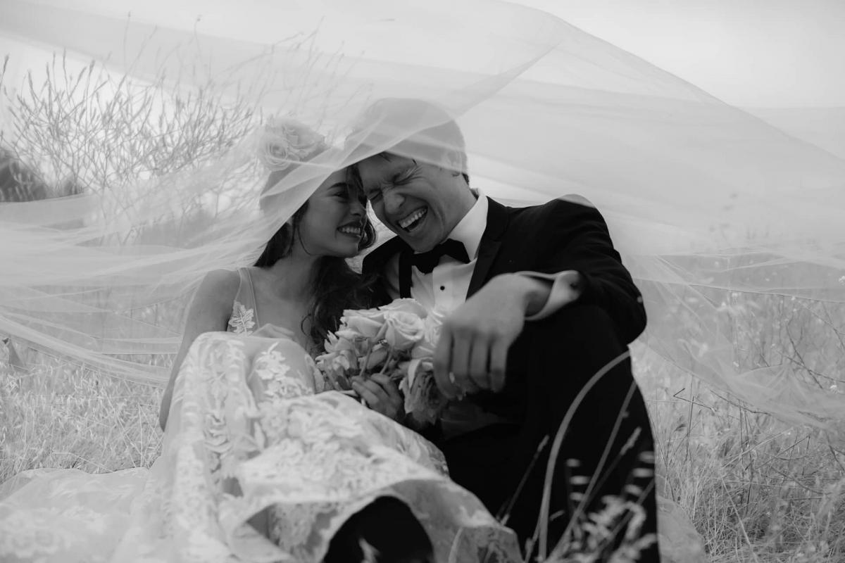 Black & White of Bride & Groom Laughing, Sitting in Field with Veil Overhead for Mika & James's Wedding