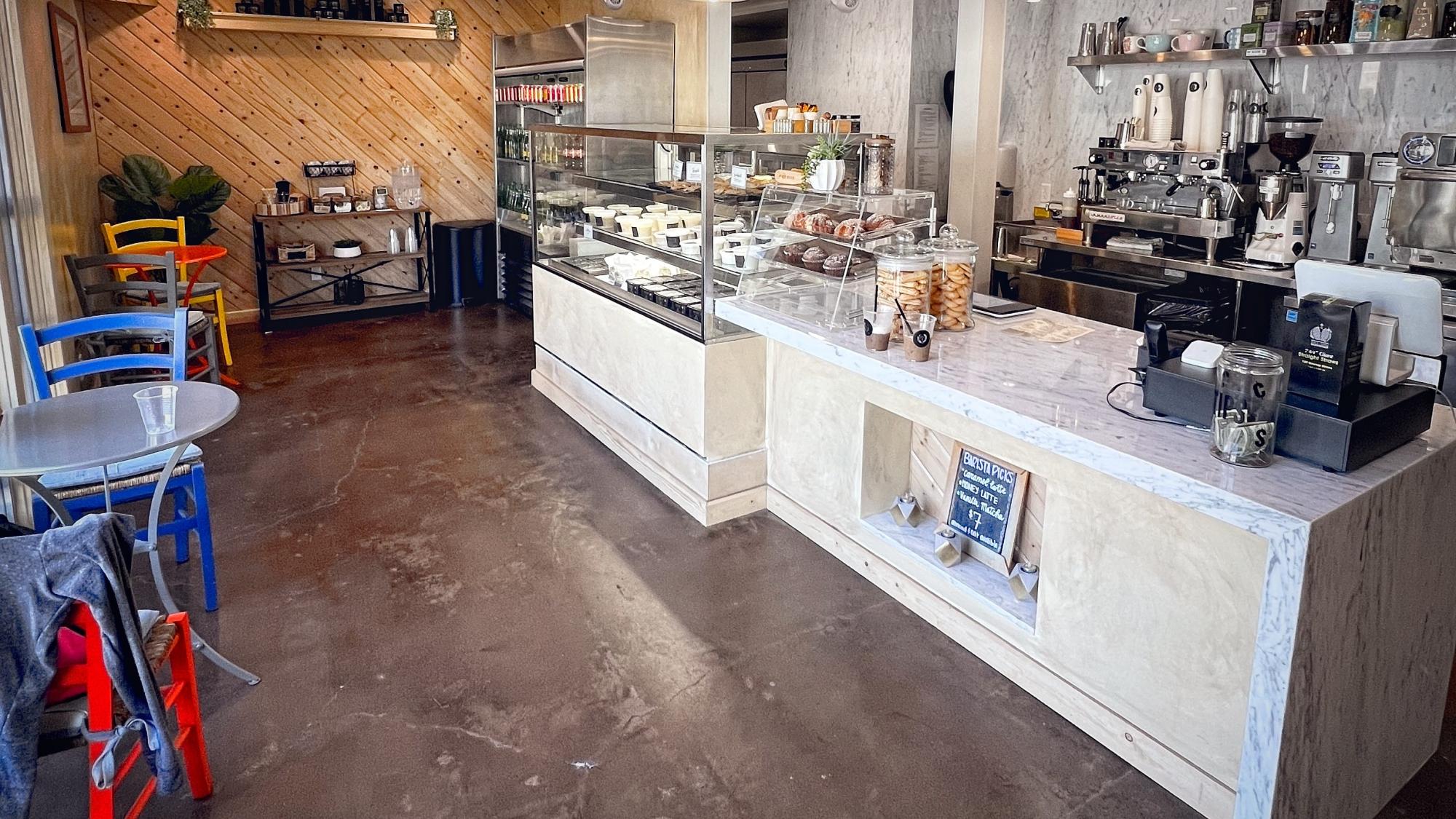A modern café interior with a marble counter, pastry display case, coffee machines, metal chairs, and wooden accents, with baked goods and drinks visible behind the counter.
