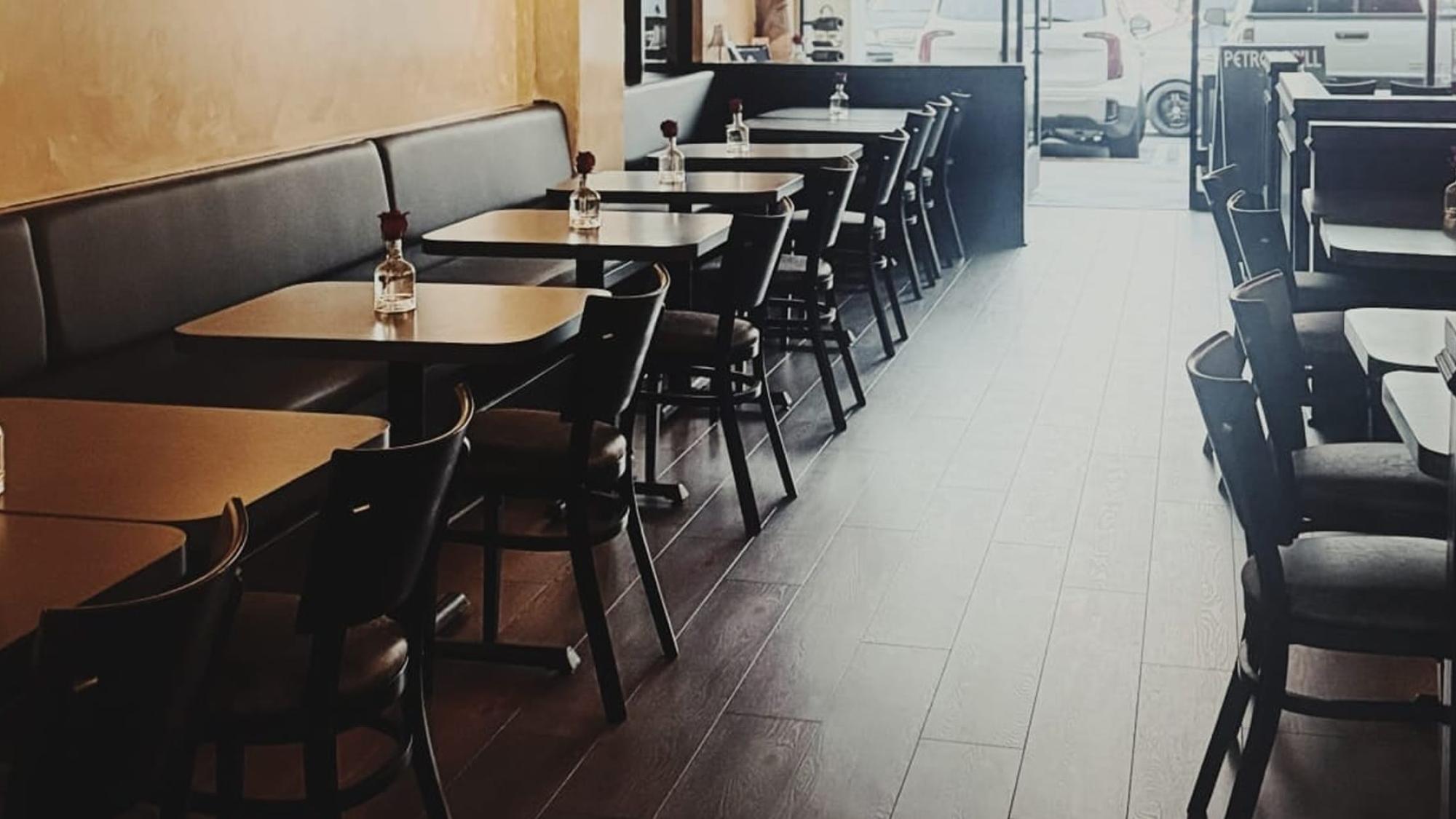 Empty restaurant with dark wooden tables and chairs, single flower vases on each table, and natural light coming through the front windows.