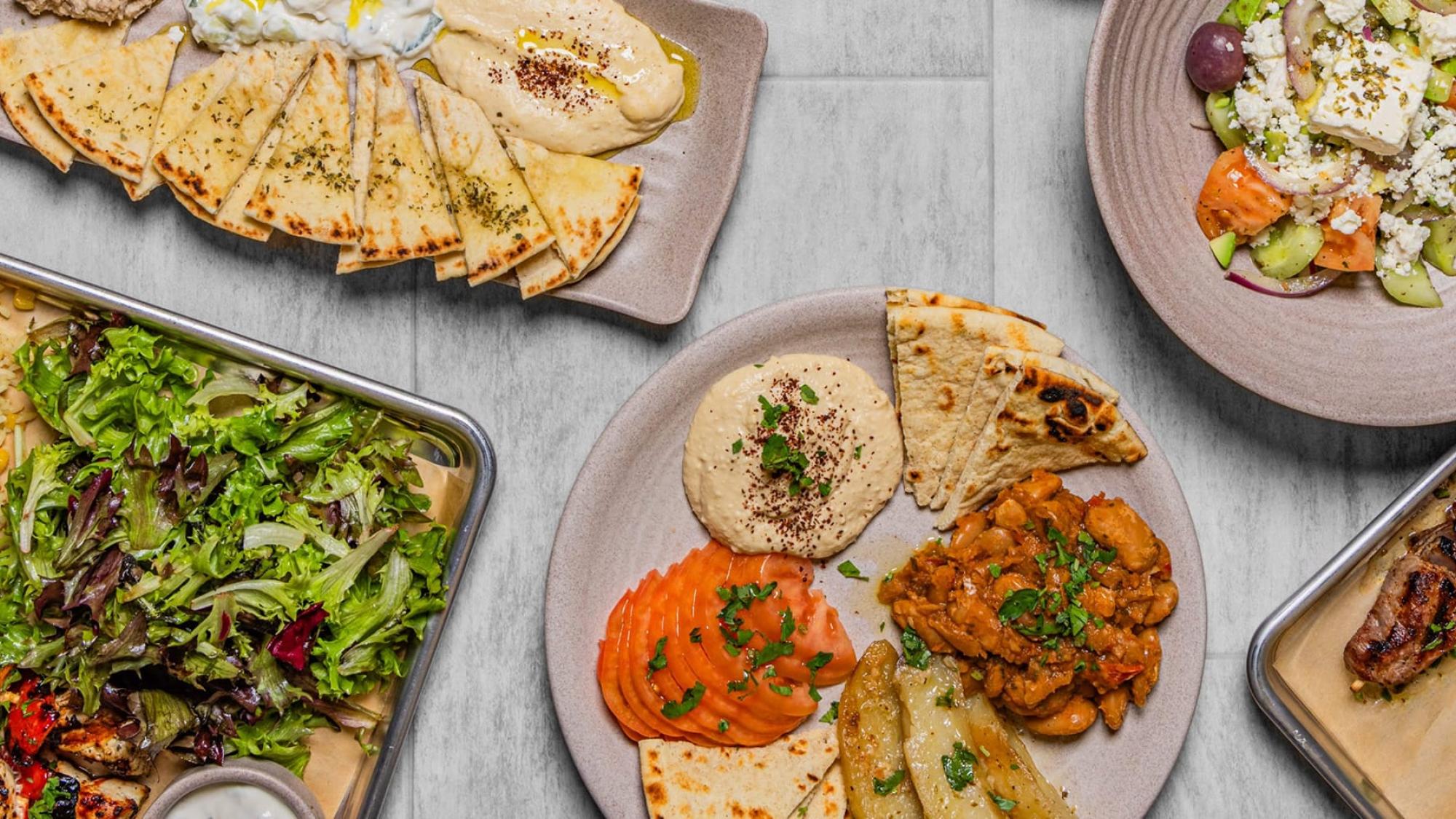 Several plates of Mediterranean food, including grilled skewers, pita bread, hummus, rice, salad, and vegetables, arranged on a light-colored tabletop.