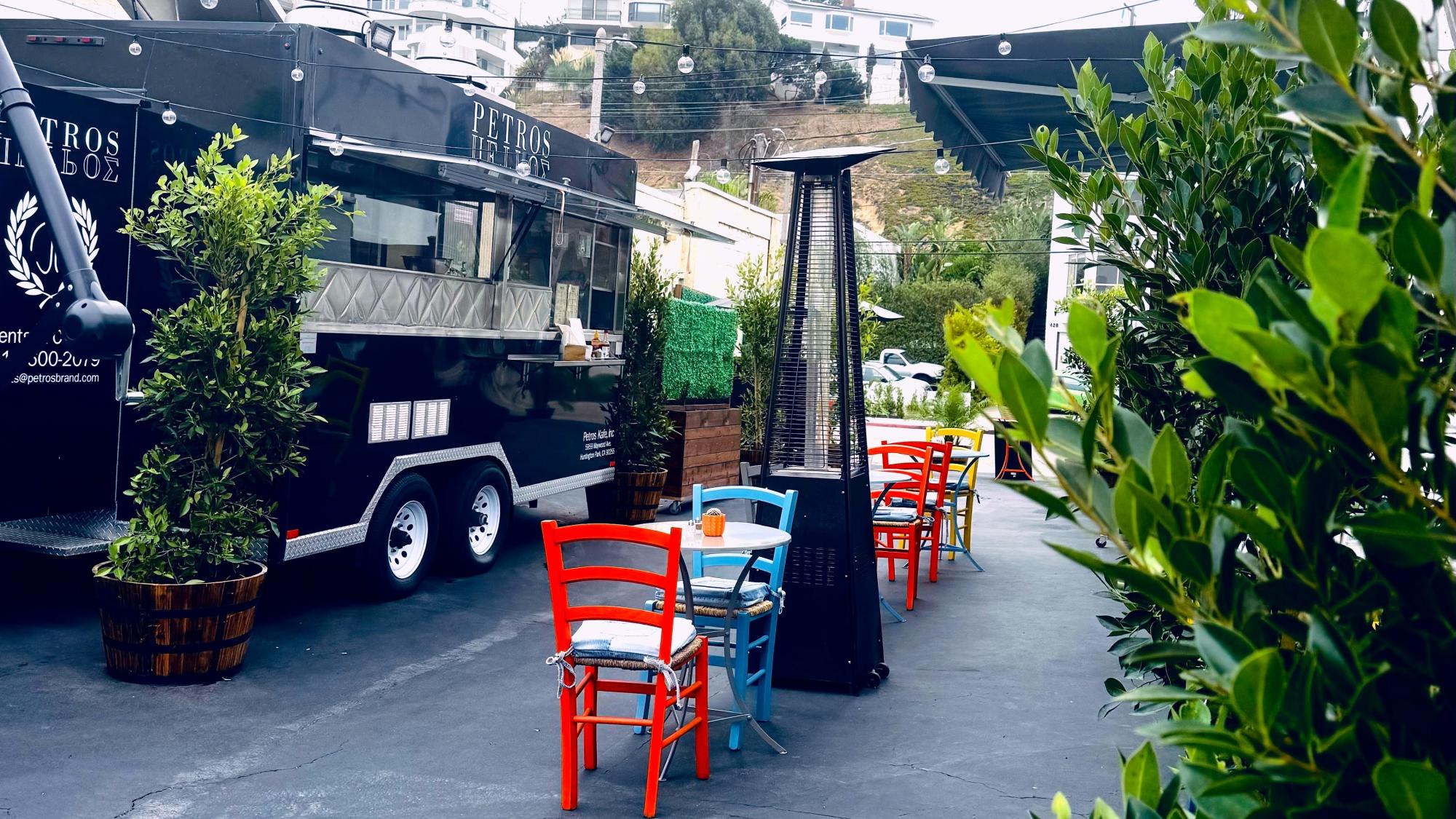 Outdoor seating area with colorful chairs and tables beside a black food truck, surrounded by potted plants and a tall outdoor heater.