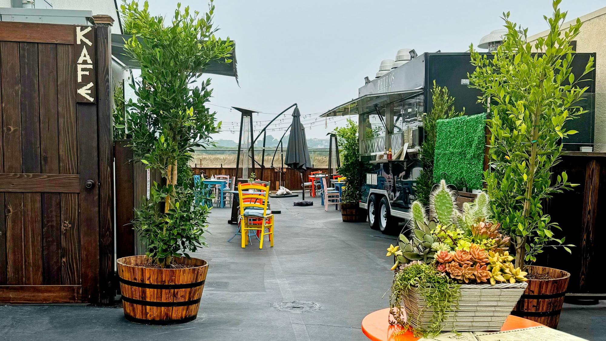 Outdoor café seating area with colorful chairs, potted plants, a food truck, and a wooden "KAFE" sign on the left gate. A flower arrangement sits on an orange table in the foreground.
