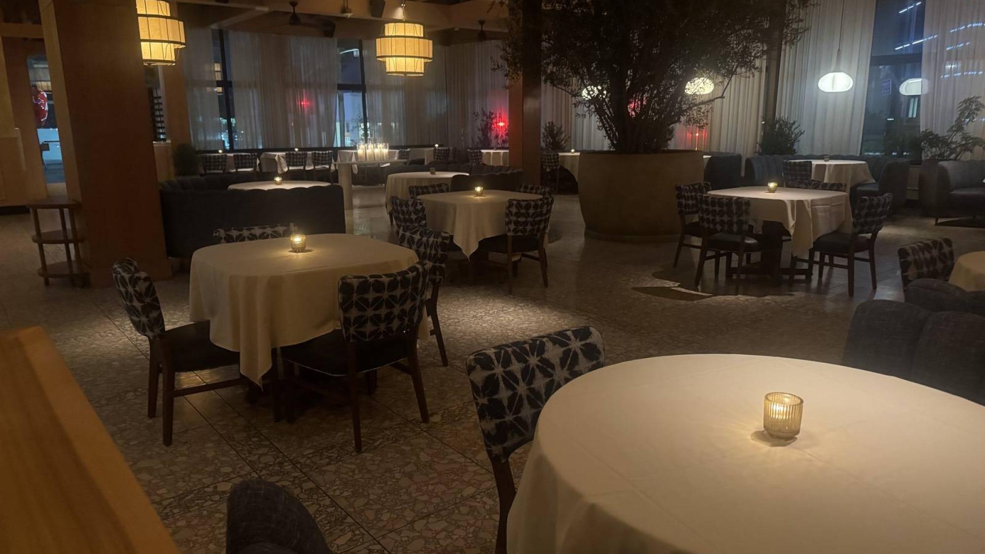 Candlelit empty dining tables with patterned chairs are arranged in a dimly lit restaurant interior.