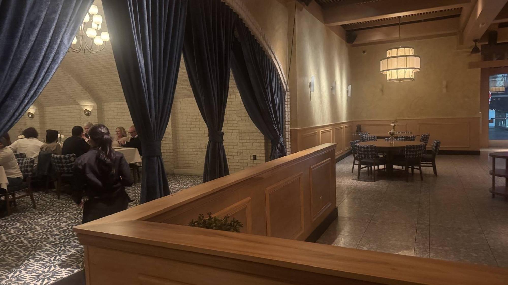 A server walks past a group of diners in a private room separated by dark blue curtains in a warmly lit restaurant.