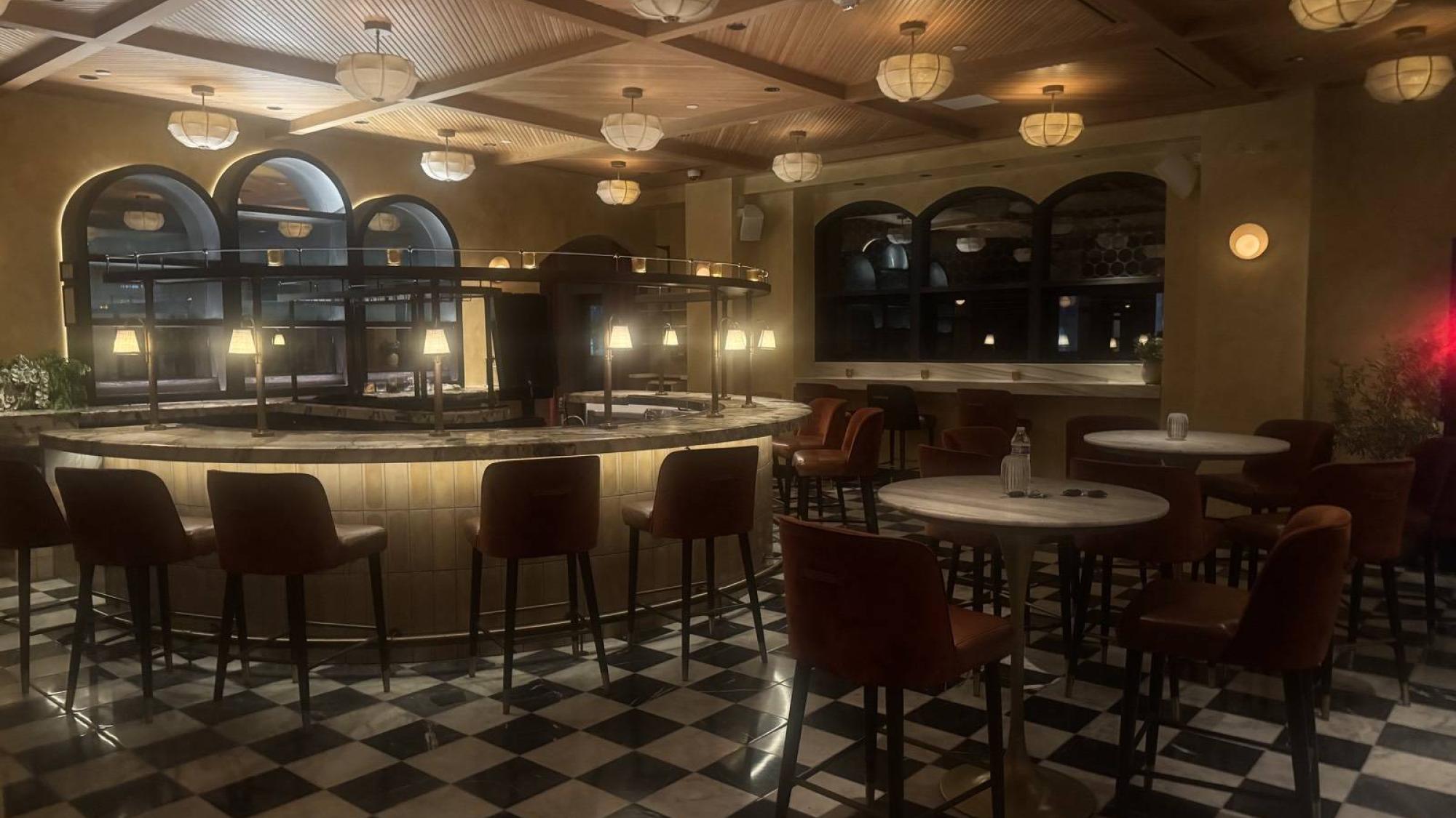 A dimly lit bar with round tables, brown chairs, and glowing pendant lights overhead.